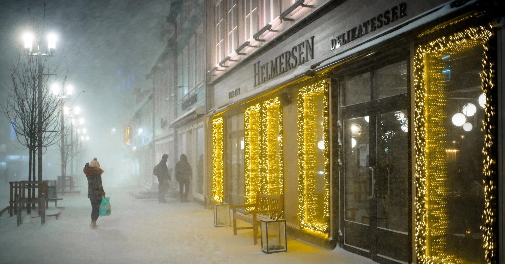 The storefront with the branded Helmersen Delikatesser sign lit up during a snowstorm