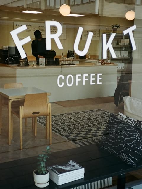 The Frukt Coffee storefront window with white lettering reading Frukt Coffee, a barista working behind the counter and a wooden chair and patterned rug visible inside