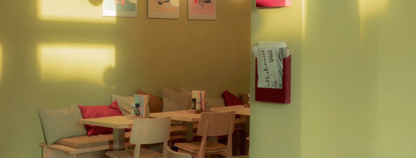 Sunlit olive green walls with framed art above a banquette set with red and beige pillows at Cupp