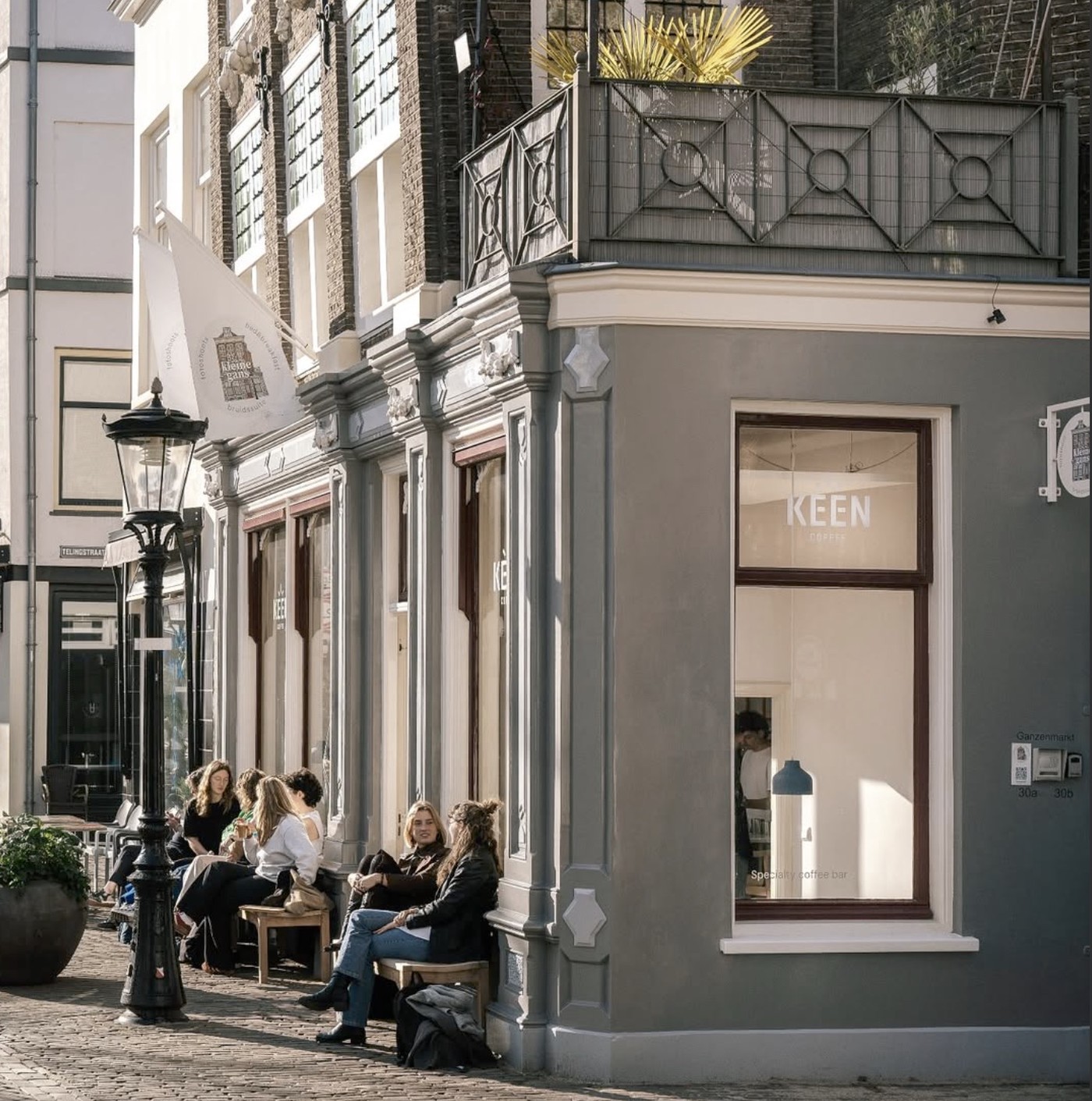 The corner storefront of Keen Coffee Bar with customers seated on a bench outside on a sunlit cobblestone street