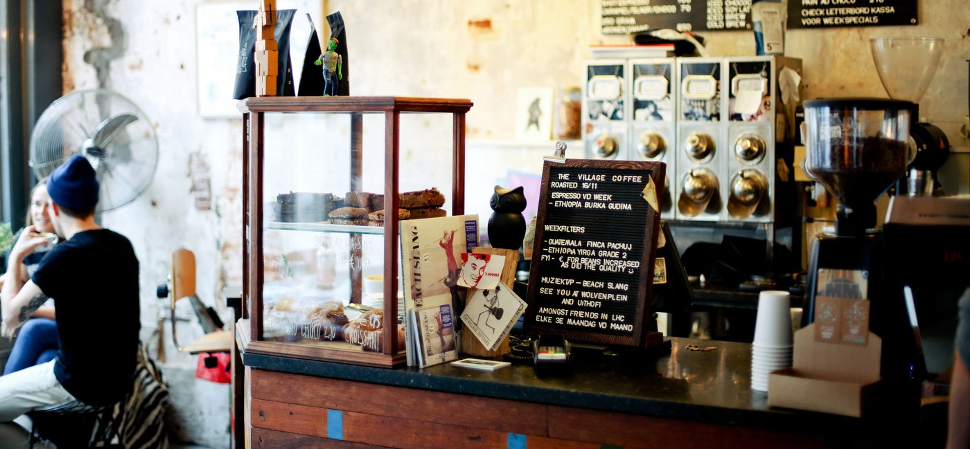 The pastry case and handwritten weekly specials board on the wooden counter at The Village Coffee & Music, with customers seated at a table in the background