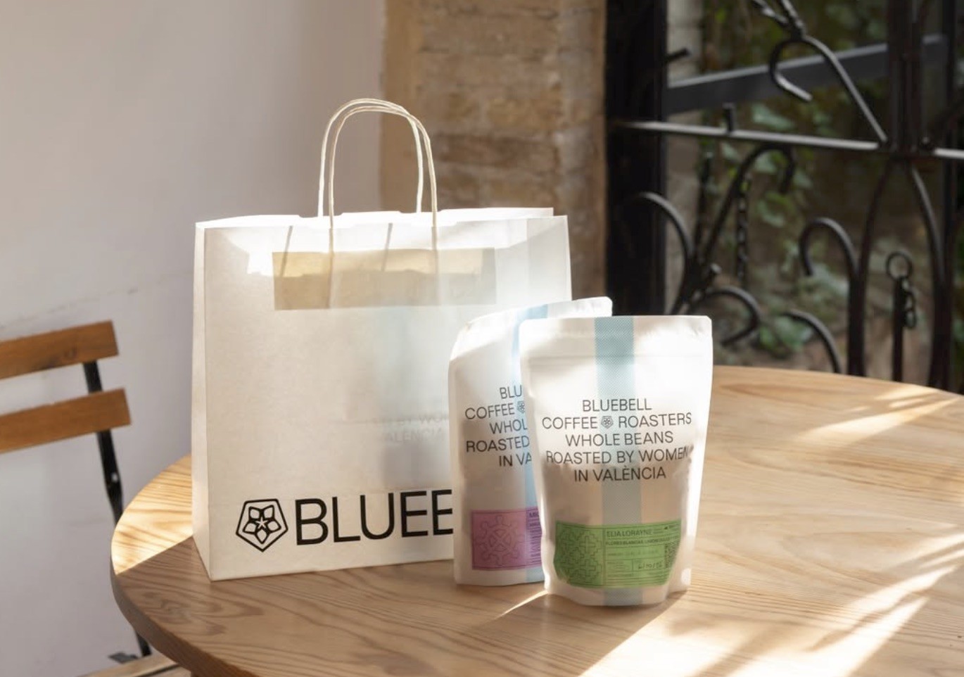 A branded paper bag and two retail coffee bags reading Roasted by Women in València on a sunlit courtyard table at Bluebell Coffee Co.