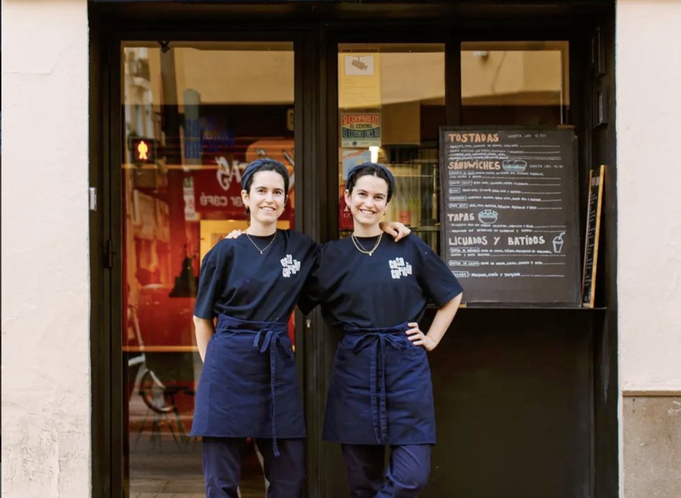 Sisters Laura and Marta Benito in matching Casa Capicúa aprons standing outside the shop beside the chalkboard menu