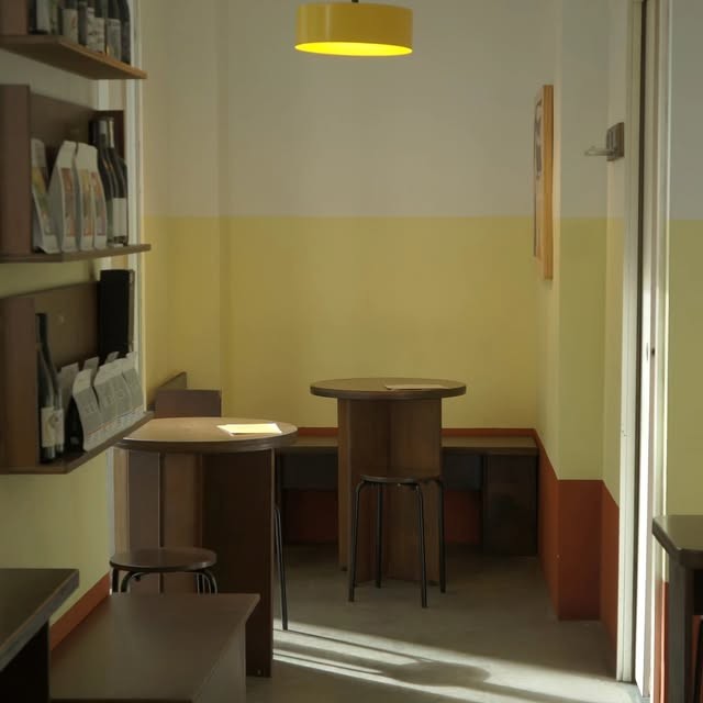 A cozy yellow walled corner of Los Picos with round wooden tables, stools, and a wine shelf lit by a pendant lamp