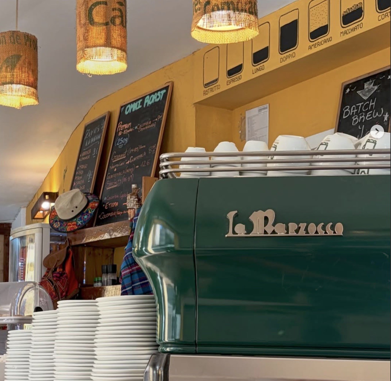 A green La Marzocco espresso machine beneath hanging burlap lamps and handwritten roast menus at Mayan Coffees