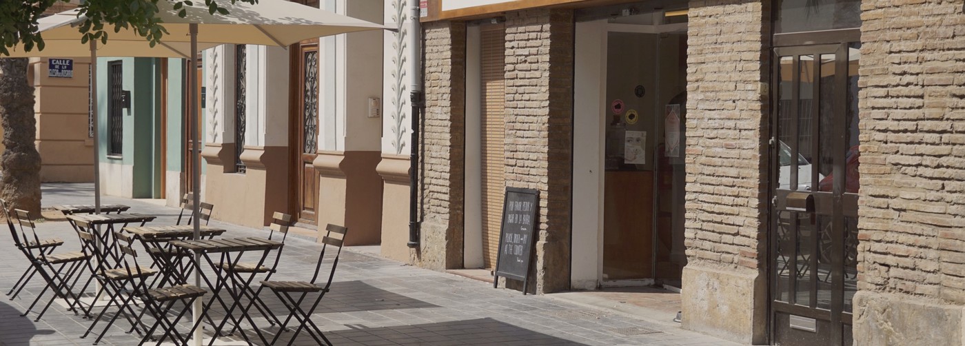 Outdoor bistro tables under an awning beside the brick entrance of TALLAT on a sunlit street