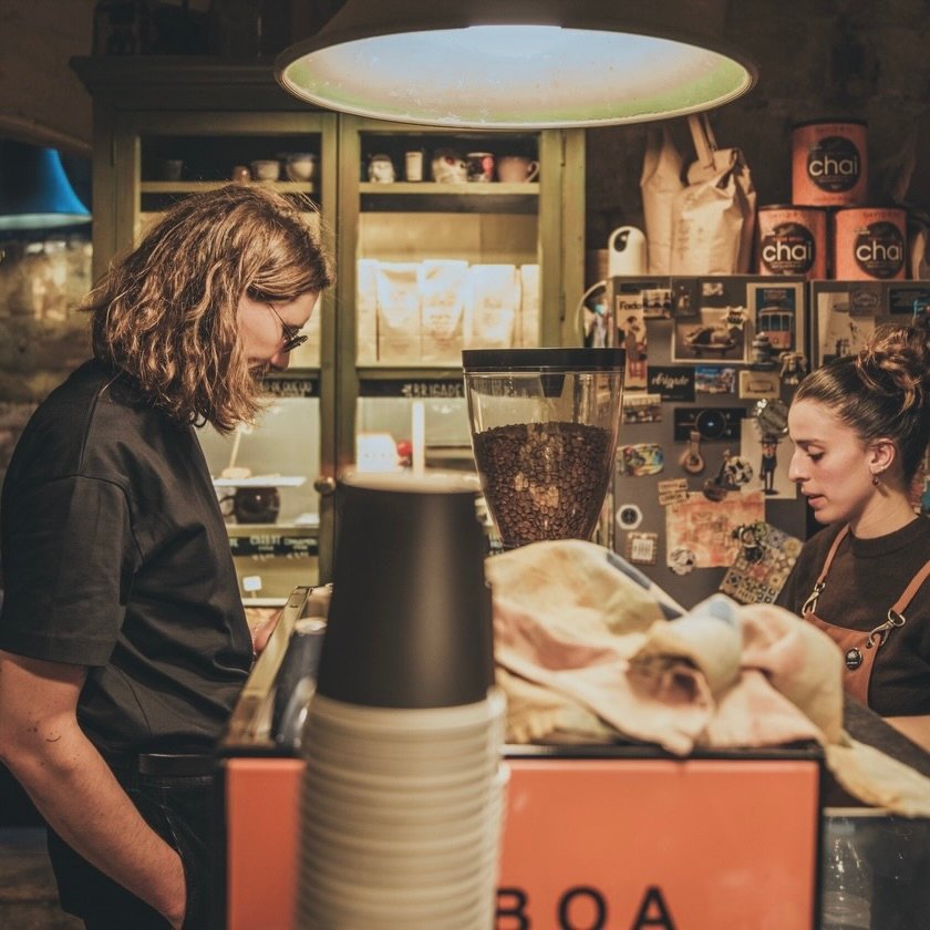 A barista working the counter at Coffee Circus Lisboa surrounded by pendant lighting and bags of chai