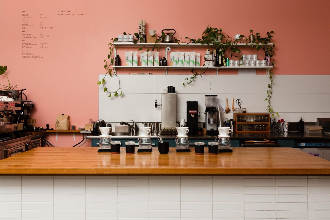 The pink walled counter at Modus Coffee Roasters, with a row of pour over drippers on the wood bar and coffee bags and mugs on the shelf above