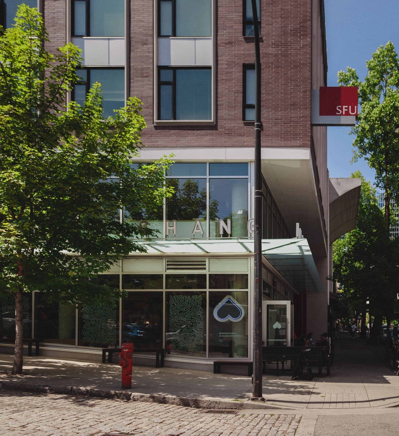 The glass fronted corner exterior of Nemesis Coffee's Gastown flagship on West Hastings Street, with a lit heart shaped sign in the window and outdoor benches along the sidewalk