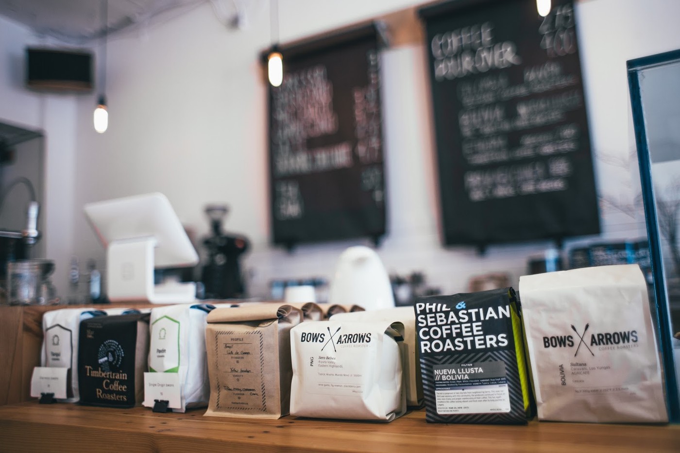 A wooden shelf at Propaganda Coffee lined with bags from Canadian roasters including Phil and Sebastian, Bows and Arrows, and Timbertrain, with chalkboard menus blurred in the background