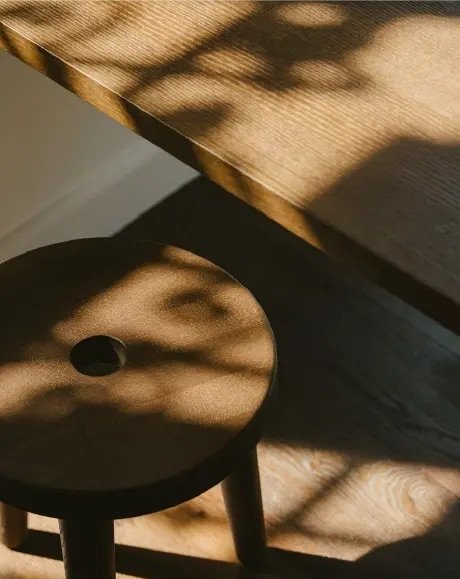 Afternoon light through a window casts leaf shaped shadows across a wooden table and stool inside the cafe