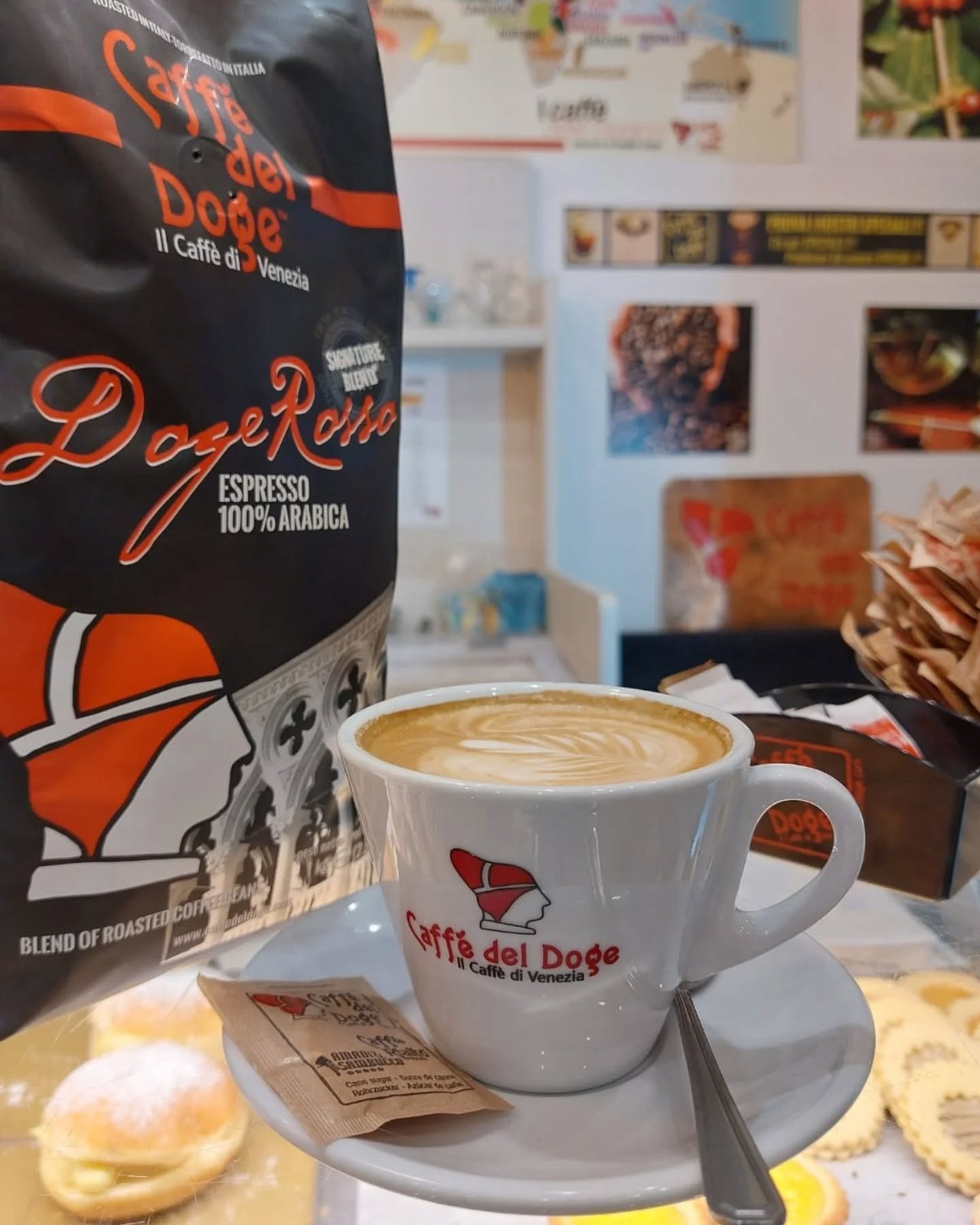 A cappuccino with latte art in a Caffè del Doge branded cup, next to a bag of Doge Rosso espresso beans and pastries