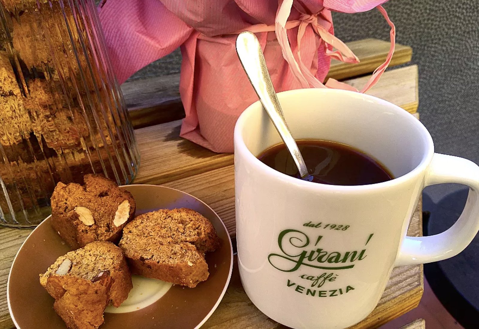 A branded Girani mug of coffee with a spoon, next to a plate of almond biscotti and a jar of roasted beans