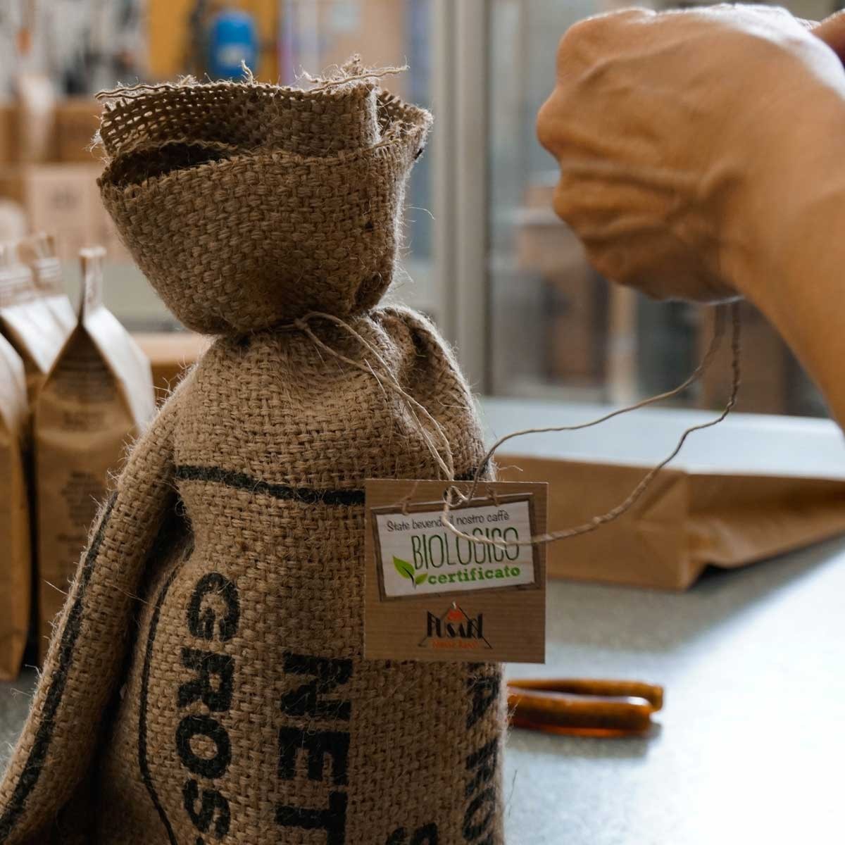A burlap coffee sack tied with string and an organic certification tag bearing the Fusari logo, a hand tying the knot