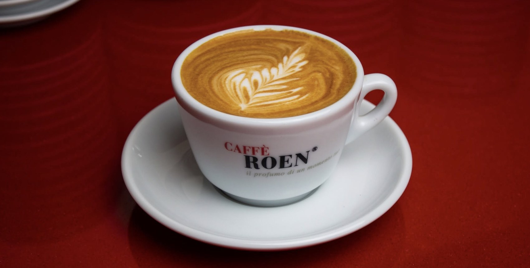 A cappuccino with latte art in a branded Caffè Roen cup and saucer on a red table