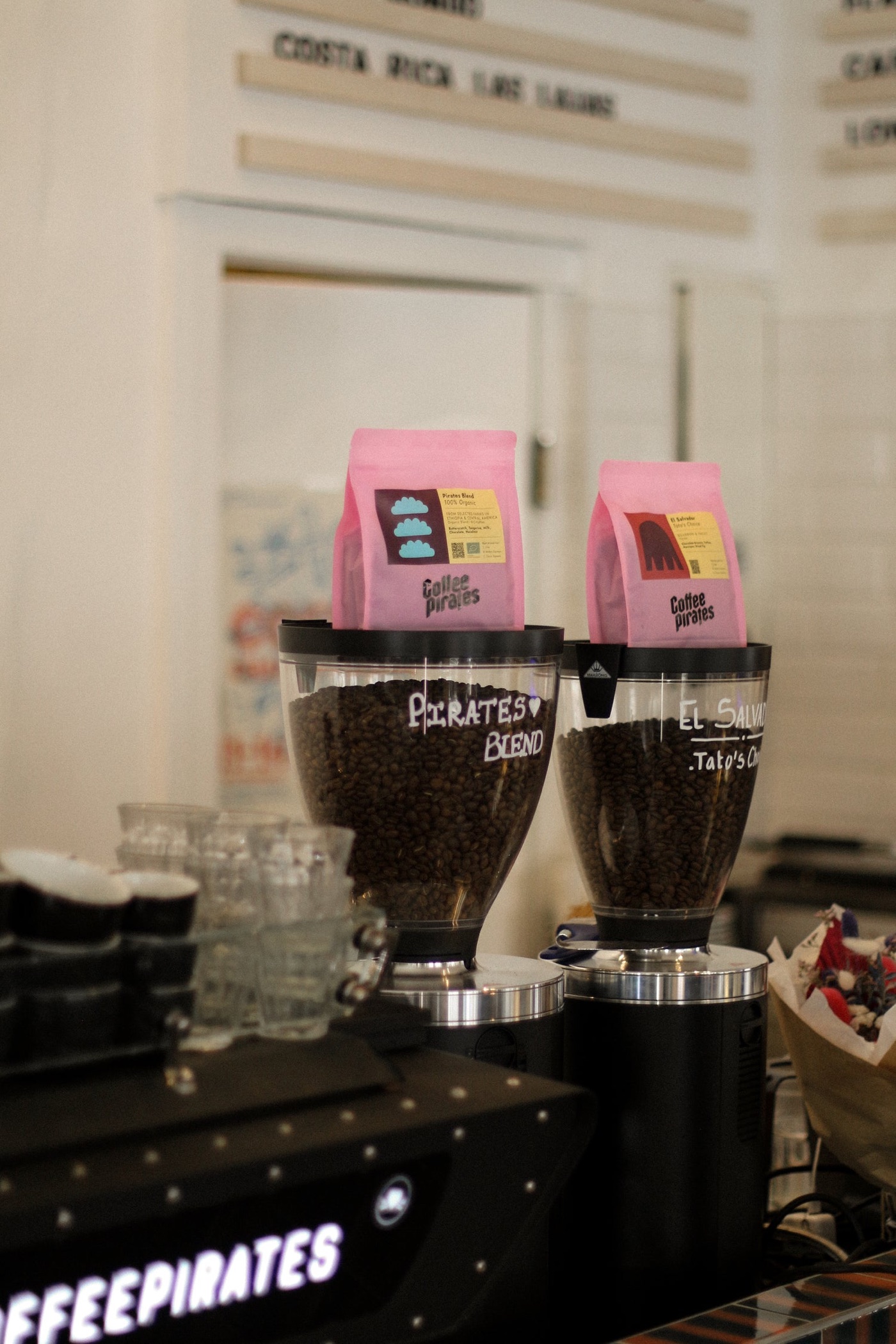 Branded Coffee Pirates bean bags atop grinder hoppers labeled Pirates Blend and El Salvador, neon sign glowing below