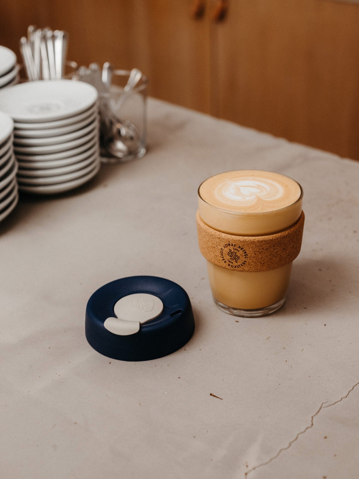 A branded Jonas Reindl reusable cup with latte art, stacked saucers in the background