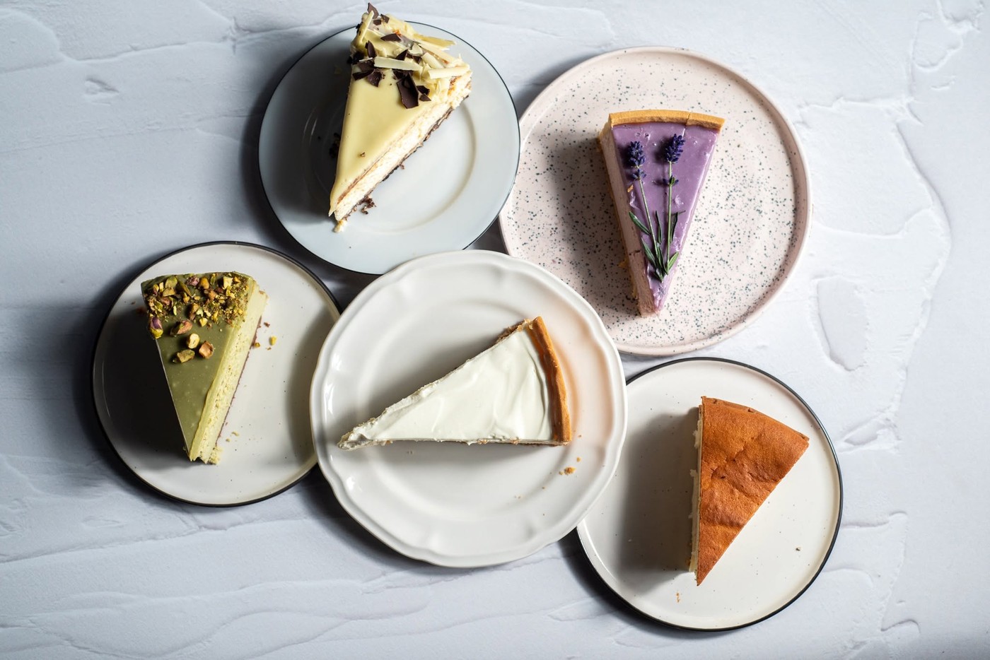 Five cake slices, including pistachio, lavender, and white chocolate flavors, arranged on mismatched plates from above at Żona Krawca