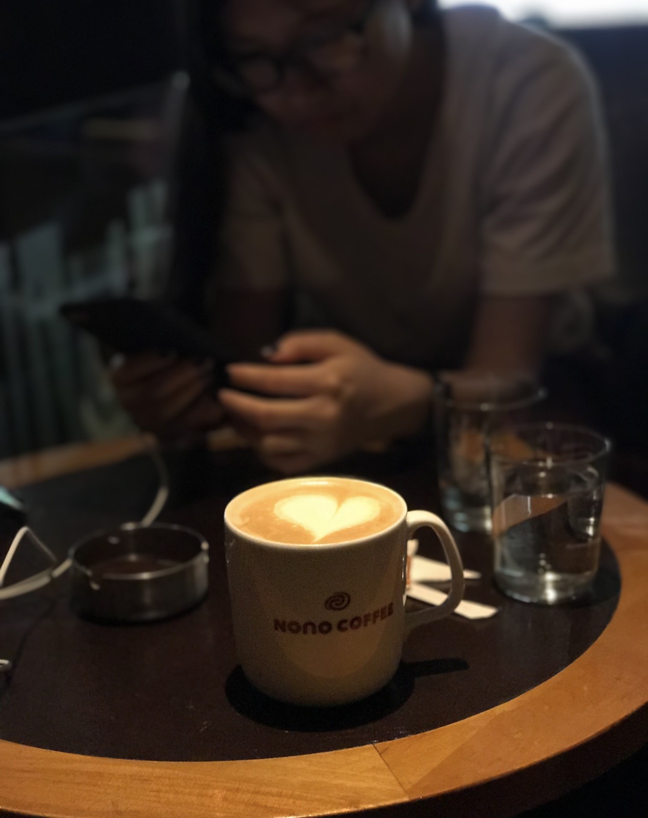 A latte with heart shaped foam art in a mug branded Nono Coffee, sitting on a dark round table