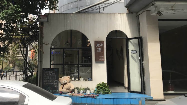 The arched white storefront with a Stand and Stare sign, a teddy bear and small plants set out by the open door