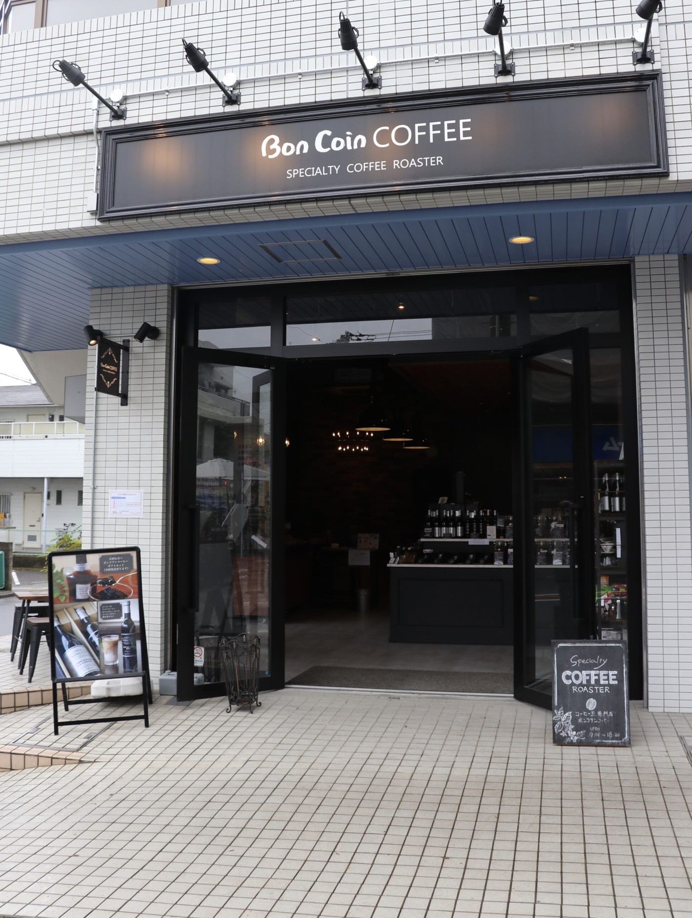 The tiled storefront of Bon Coin Coffee with its dark signboard, an open terrace with stools and small tables in front of the glass doors