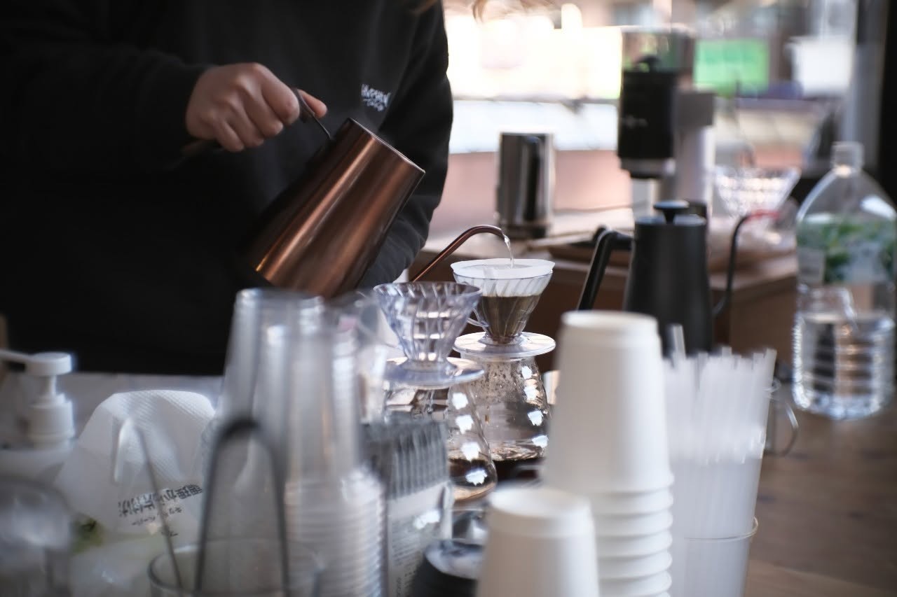 A barista pouring hot water over a V60 dripper at the counter, with stacked cups, straws, and brewing gear lined up on the bar