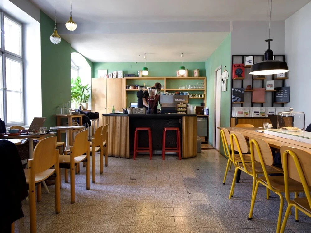 The green walled interior of Cogito Coffee Deželićeva, with wooden chairs and yellow chairs around a communal table
