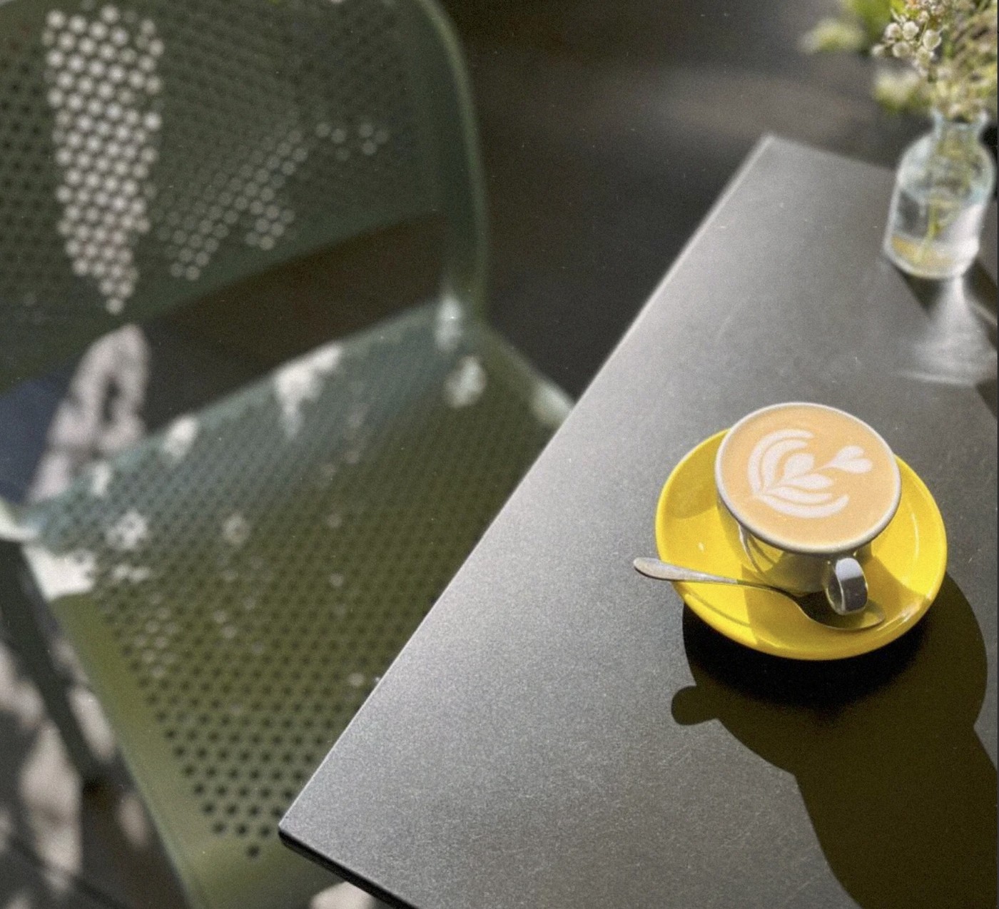 A cappuccino with leaf latte art in a yellow cup and saucer on a dark table beside a perforated metal chair