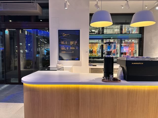 The wood-fronted counter at Commercial - The Project at night, espresso machine and menu screen lit against the storefront glass