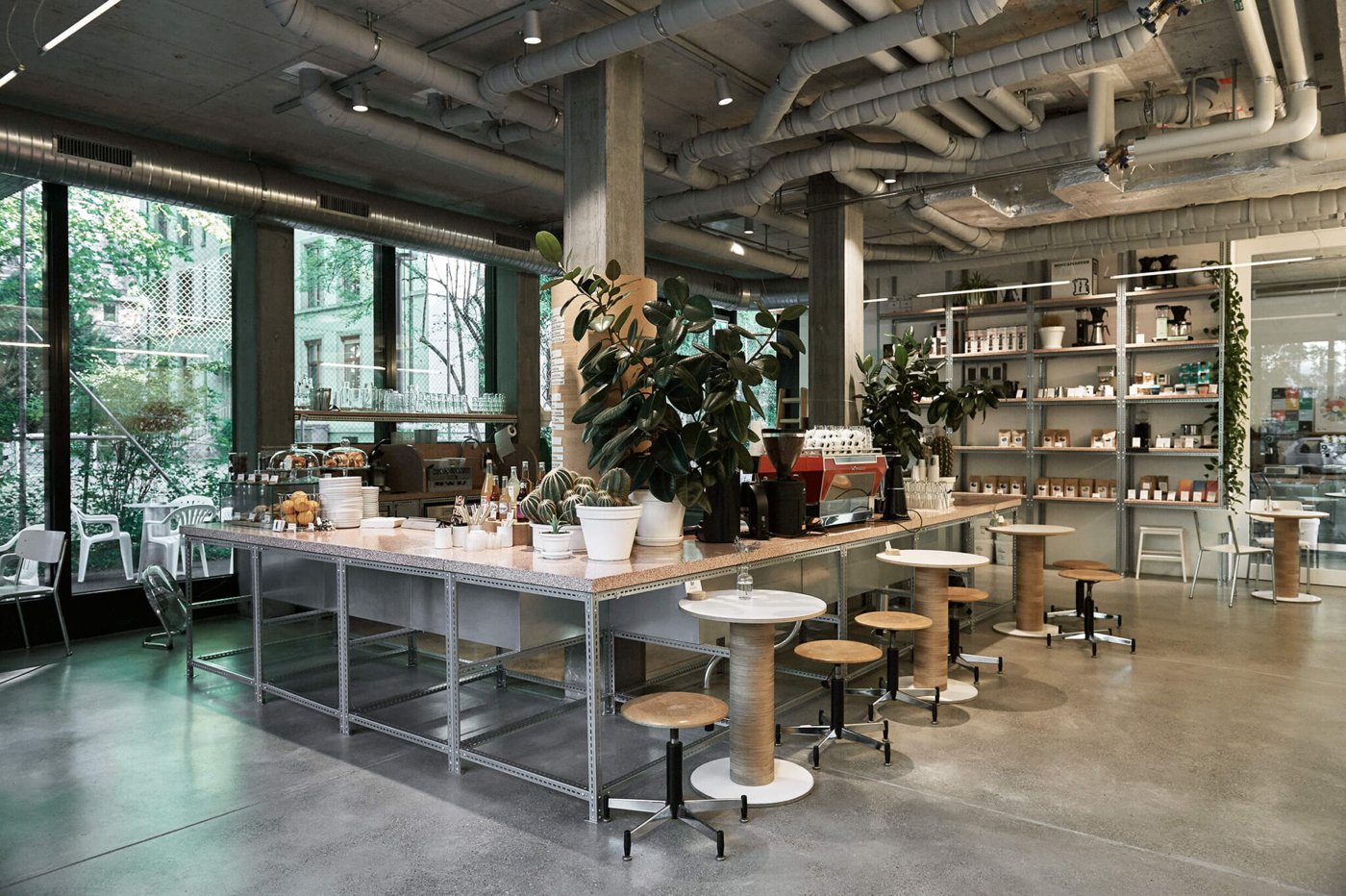 The industrial interior of Miro Manufactura de Cafe, concrete pillars, exposed piping, and a long communal counter with the roaster visible